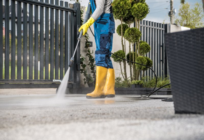 A worker in boots & overalls cleans a driveway in Phoenix, AZ, showcasing summer cleaning services.